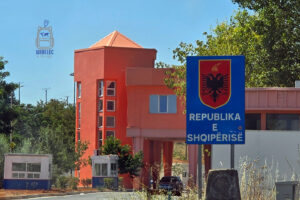 Przejście graniczne prowadzące do Albanii, z dużą niebieską tablicą informacyjną z napisem „Republika e Shqipërisë”, oznaczającym Republikę Albanii. Nad napisem widnieje czerwony herb z czarnym dwugłowym orłem, symbolem narodowym Albanii. W tle znajduje się charakterystyczny budynek kontroli granicznej w pomarańczowym kolorze, niewielkie budki strażnicze oraz droga prowadząca przez przejście. Słoneczna pogoda i otaczająca zieleń towarzyszą kolejnemu etapowi bałkańskiej podróży.