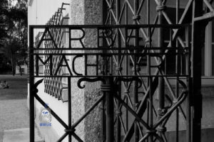 Czarno-białe zdjęcie przedstawiające zbliżenie na metalową bramę byłego nazistowskiego obozu koncentracyjnego Dachau w Niemczech, z cynicznym napisem „Arbeit macht frei” („Praca czyni wolnym”). Żelazna konstrukcja z charakterystycznymi geometrycznymi zdobieniami wypełnia większość kadru. W tle widoczny jest fragment budynku Jourhaus oraz teren dawnego obozu. Monochromatyczna fotografia podkreśla surowość miejsca i stanowi przypomnienie o cierpieniu więźniów oraz zbrodniach nazistowskiego systemu obozów koncentracyjnych.