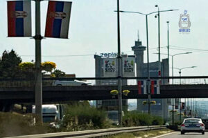 Widok na Belgrad w Serbii z charakterystyczną wieżą Genex, znaną jako Zachodnia Brama Belgradu, w tle. Na pierwszym planie ruchliwa droga, wiadukt i serbskie flagi, a na wieży widoczne reklamy wydarzenia EXPO 2027