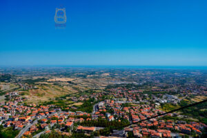 Rozległa panorama Borgo Maggiore i okolic San Marino widziana z wysokości Monte Titano. Na pierwszym planie rozciąga się zabudowa miasta z charakterystycznymi czerwonymi dachami, krętymi ulicami i budynkami otoczonymi zielenią. Dalej widoczne są łagodne wzgórza, pola uprawne i niewielkie miejscowości rozsiane po krajobrazie regionu Emilia-Romania. Na odległym horyzoncie można dostrzec błękitne wody Morza Adriatyckiego, niemal zlewające się z bezchmurnym niebem.