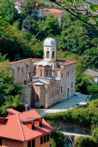Widok z góry na zabytkową Cerkiew Chrystusa Zbawiciela (Crkva Svetog Spasa) w Prizren w Kosowie. Niewielka średniowieczna świątynia prawosławna z charakterystyczną ceglaną kopułą otoczona jest ruinami większej budowli o kamiennych murach i dzwonnicy zwieńczonej jasną kopułą. Cerkiew położona jest na zielonym zboczu wzgórza, wśród gęstych drzew i tradycyjnej zabudowy z czerwonymi dachami. Zdjęcie wykonane w słoneczny dzień ukazuje kontrast między historyczną architekturą sakralną a otaczającą ją przyrodą.