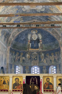 Wnętrze zabytkowej cerkwi św. Zofii w Ochrydzie w Macedonii Północnej, z monumentalnymi bizantyjskimi freskami zdobiącymi apsydę ołtarzową i sklepienie świątyni. W centralnej części górnego malowidła przedstawiona jest Matka Boska z Dzieciątkiem, otoczona złotą aureolą, a poniżej znajdują się postacie świętych i sceny liturgiczne. Freski utrzymane są w przygaszonych odcieniach błękitu, szarości i złota, nosząc widoczne ślady upływu czasu. W dolnej części fotografii znajduje się ikonostas z barwnymi ikonami Matki Boskiej i Chrystusa, ozdobnymi kolumnami oraz centralnym wejściem do prezbiterium. Przez charakterystyczne okna z okrągłymi otworami wpada naturalne światło. Drewniane belki przecinające sklepienie podkreślają historyczny charakter wnętrza. Fotografia ukazuje bogactwo średniowiecznej sztuki sakralnej i bizantyjskiego dziedzictwa Ochrydy.