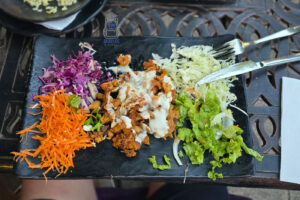 Döner kebab podany na czarnym, prostokątnym talerzu w jednej z restauracji w Prizren w Kosowie. W centralnej części talerza znajdują się kawałki przypieczonego mięsa polane białym sosem, otoczone kolorowymi dodatkami: surówką z czerwonej i białej kapusty, startą marchewką oraz świeżą zieloną sałatą. Obok potrawy leżą nóż i widelec, a całość ustawiona jest na ozdobnym metalowym stoliku restauracyjnym. Zdjęcie przedstawia jeden z popularnych smaków bałkańskiej kuchni ulicznej.