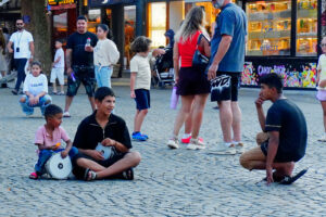 Dwóch chłopców siedzących na brukowanym placu w historycznym centrum Prizren w Kosowie, z niewielkimi bębnami trzymanymi na kolanach. Starszy chłopiec uśmiecha się, trzymając instrument, a młodszy siedzi tuż obok niego. Po prawej stronie przykucnął nastolatek obserwujący scenę. W tle widać spacerujących mieszkańców i turystów, rodziny z dziećmi oraz kolorowo oświetlone witryny sklepów, w tym sklep ze słodyczami. Fotografia oddaje atmosferę codziennego życia i wieczornego gwaru ulic Prizren.