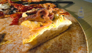 Zbliżenie na kawałek włoskiej focacci carbonara podany na ceramicznym talerzu. Puszyste, dobrze wyrośnięte ciasto o chrupiącej, złocistej skórce pokryte jest kremowym, żółtym sosem oraz kawałkami rumianego, chrupiącego boczku. Widoczny przekrój ukazuje lekką, porowatą strukturę pieczywa. W tle znajdują się pozostałe kawałki pizzy z pomidorami i serem.