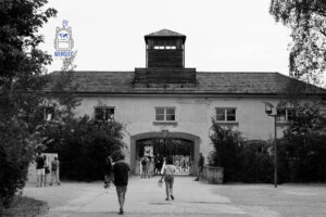 Czarno-białe zdjęcie przedstawiające Jourhaus, historyczny budynek bramy wejściowej do byłego nazistowskiego obozu koncentracyjnego Dachau w Niemczech. Nad centralnym przejściem z metalową bramą wznosi się charakterystyczna drewniana wieżyczka strażnicza. Przed budynkiem widoczni są zwiedzający zmierzający w kierunku wejścia na teren miejsca pamięci. Surowa architektura, otaczające drzewa i monochromatyczna kolorystyka podkreślają powagę oraz tragiczne znaczenie historyczne tego miejsca.