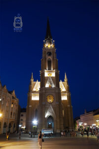 Neogotycki kościół Imienia Maryi przy Placu Wolności (Trg slobode) w Nowym Sadzie w Serbii, sfotografowany nocą. Wysoka wieża zegarowa, strzeliste detale architektoniczne i rozświetlona rozeta wyróżniają się na tle granatowego nieba. Przed kościołem spacerują mieszkańcy i turyści.