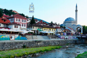 Panorama zabytkowego centrum Prizren w Kosowie, z monumentalnym meczetem Sinana Paszy, którego charakterystyczna niebieskawa kopuła i wysoki, smukły minaret górują nad staromiejską zabudową. Wzdłuż rzeki Bistricy ciągną się kolorowe kamienice z czerwonymi dachami, restauracje i kawiarnie z ogródkami pod białymi parasolami. Na pierwszym planie widoczne jest kamieniste koryto rzeki oraz kamienne nabrzeże. W tle zielone wzgórza otaczające miasto. Fotografia ukazuje wyjątkową atmosferę Prizren, łączącego osmańską architekturę z tętniącym życiem współczesnego miasta.