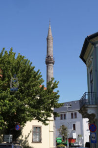 Zabytkowy turecki minaret w Egerze na Węgrzech, wznoszący się ponad dachami kamienic i zielonymi drzewami. Na wąskim balkonie widokowym stoją turyści, a smukła kamienna wieża odcina się od błękitnego nieba.