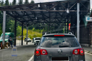 Przejście graniczne między Czarnogórą a Kosowem, widziane zza przedniej szyby samochodu. Na pierwszym planie stoi szary Volkswagen Passat z zapalonymi światłami hamowania, oczekujący na odprawę. Nad drogą znajduje się duża metalowa wiata, pod którą odbywa się kontrola graniczna. W tle widać kolejne samochody, zabudowania przejścia oraz zielone, zalesione wzgórza. Zdjęcie dokumentuje moment przekraczania granicy podczas samochodowej podróży przez Bałkany.
