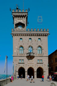 Palazzo Pubblico, czyli Pałac Publiczny w San Marino, stojący przy Piazza della Libertà. Monumentalny neogotycki budynek z jasnego kamienia wyróżnia się wieżą zegarową zwieńczoną blankami i dzwonnicą. Fasada ozdobiona jest ostrołukowymi oknami, herbami oraz balkonem z flagą San Marino. Na parterze znajdują się trzy arkadowe wejścia, a przed pałacem spacerują turyści. W tle rozciąga się intensywnie błękitne niebo.
