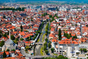 Rozległa panorama Prizren w Kosowie, widziana z górującej nad miastem twierdzy Kalaja. Na pierwszym planie znajduje się zabytkowy meczet Sinana Paszy z charakterystyczną kopułą i wysokim minaretem. Przez centrum miasta przepływa kamienista rzeka Bistrica, nad którą przerzucono zabytkowy kamienny most. Gęsta zabudowa z czerwonymi dachami, liczne minarety i zielone drzewa tworzą malowniczy krajobraz historycznej części Prizren. W oddali widoczne są nowoczesne budynki, rozległa dolina i otaczające miasto góry.