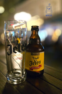 Butelka serbskiego piwa Jelen Svetlo Pivo z charakterystyczną żółtą etykietą i wizerunkiem jelenia, stojąca obok pustej szklanki z logo marki na drewnianym stoliku. W tle rozmyte, ciepłe światła wieczornego lokalu.