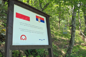 Tablica informacyjna przy Jaskini Lodowej (Ledena Pećina) w rezerwacie przyrody Uvac w Serbii. Na tablicy umieszczono flagi Polski i Serbii oraz informację, że zagospodarowanie jaskini zostało współfinansowane ze środków Ministerstwa Spraw Zagranicznych RP w ramach programu Polska Pomoc. Tablica stoi w otoczeniu zielonego lasu