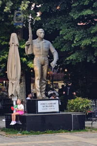 Pomnik Ismeta Jashariego, znanego jako „Kumanova”, dowódcy Armii Wyzwolenia Kosowa (UÇK), znajdujący się w Prizren. Monument przedstawia stojącego mężczyznę w wojskowym stroju, trzymającego karabin w prawej ręce. Na czarnym cokole umieszczono tablicę z nazwiskiem oraz datami życia: 16 kwietnia 1967 – 25 sierpnia 1998. U podstawy pomnika siedzi mała dziewczynka, a w tle widać ogródki kawiarniane i rozłożyste, zielone drzewa. Zdjęcie ukazuje połączenie pamięci o historii Kosowa z codziennym życiem miasta.