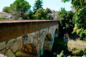 Zabytkowy Ponte Consolare w Savignano sul Rubicone we Włoszech, kamienny most o rzymskich korzeniach, przerzucony nad rzeką Rubikon. Fotografia przedstawia masywne kamienne łuki mostu, zbudowane z dużych jasnych bloków i zwieńczone ceglaną balustradą. W tle widoczne są drzewa, bujna roślinność oraz zabudowania włoskiego miasteczka. Most stanowi świadectwo rzymskiego dziedzictwa regionu i nawiązuje do historii Juliusza Cezara.