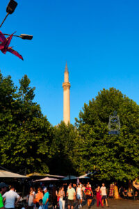 Wysoki, smukły minaret zabytkowego meczetu Sinana Paszy w Prizren w Kosowie, wznoszący się ponad koronami zielonych drzew na tle intensywnie błękitnego nieba. Kamienna wieża oświetlona ciepłym światłem popołudniowego słońca dominuje nad ruchliwym placem, na którym spacerują mieszkańcy i turyści. Wśród drzew widoczne są ogródki restauracyjne z parasolami, a po lewej stronie fragmenty czerwonych flag. Fotografia ukazuje codzienne życie historycznego centrum Prizren oraz charakterystyczne elementy jego osmańskiej architektury.