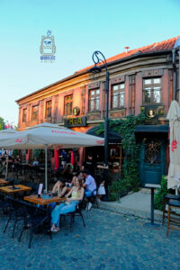 Urokliwa uliczka na starym mieście w Prizren w Kosowie, z tradycyjnym piętrowym budynkiem o drewnianej elewacji, ozdobnych oknach i czerwonym dachu. Na parterze znajdują się kawiarnia THEA oraz wejście do hotelu, otoczone zielonym bluszczem. Przed budynkiem, pod dużymi parasolami, mieszkańcy i turyści odpoczywają przy stolikach ustawionych na brukowanym deptaku. Ciepłe światło zachodzącego słońca podkreśla architekturę i przytulną atmosferę historycznego centrum Prizren.