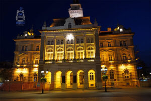 Bogato zdobiona fasada neorenesansowego ratusza w Nowym Sadzie w Serbii, sfotografowana nocą. Monumentalny budynek z wieżą zegarową, kolumnami i arkadami rozświetlają złote iluminacje, kontrastujące z ciemnogranatowym niebem