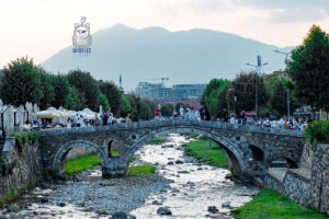 Zabytkowy Kamienny Most (Ura e Gurit) nad rzeką Bistricą w centrum Prizren w Kosowie. Charakterystyczny kamienny most z trzema łukami łączy oba brzegi płytkiej, kamienistej rzeki. Po moście spacerują mieszkańcy i turyści, a wzdłuż brzegów rozciągają się ogródki restauracyjne i aleje drzew. W tle widoczne są zabudowania miasta, smukły minaret oraz majestatyczne góry, tworzące malowniczą panoramę Prizren.