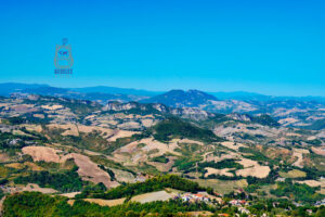 Rozległa panorama górzystego krajobrazu wokół San Marino, widziana ze szczytu Monte Titano. Łagodne wzgórza Apeninów pokrywają złociste pola uprawne, zielone lasy i niewielkie miejscowości z charakterystycznymi czerwonymi dachami. W oddali wznoszą się skaliste grzbiety i wyższe szczyty górskie, tworzące malowniczą panoramę na tle intensywnie błękitnego, bezchmurnego nieba.