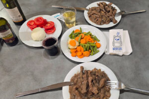 Tradycyjny włoski posiłek ze straccetti di manzo al sugo, czyli cienkich kawałków wołowiny podanych w gęstym, brązowym sosie. Na stole stoją dwa talerze z mięsem, półmisek gotowanych warzyw z marchewką, zieloną fasolką i kalafiorem oraz talerz ze świeżymi pomidorami i kulą mozzarelli. Posiłkowi towarzyszą plastikowe kubeczki z czerwonym i białym winem oraz butelki włoskiego wina. Całość podana jest na prostym, szarym blacie, tworząc obraz nieskomplikowanej, sycącej włoskiej kolacji.