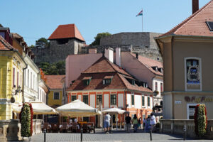Plac Istvána Dobó w Egerze na Węgrzech. Kolorowe kamienice z czerwonymi dachami, ogródki kawiarniane i spacerujący turyści. W tle kamienne mury zamku Egri Vár z powiewającą węgierską flagą