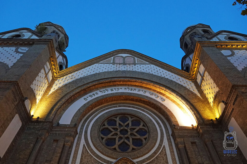 Monumentalna fasada synagogi w Nowym Sadzie w Serbii, sfotografowana z dołu o zmierzchu. Charakterystyczne ceglane wieże, duże okrągłe okno z rozetą i hebrajski napis nad wejściem. Ciepłe oświetlenie podkreśla detale architektoniczne na tle błękitnego nieba.