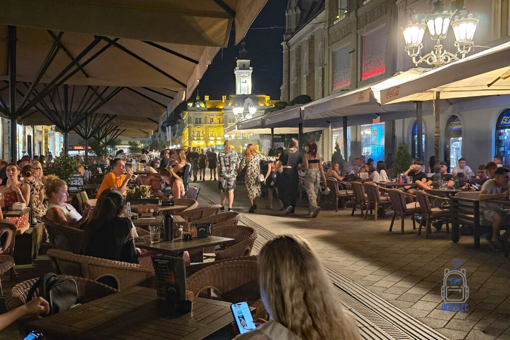 Tętniąca życiem ulica w centrum Nowego Sadu w Serbii podczas letniego wieczoru. Tłumy spacerowiczów i goście wypełniają ogródki restauracyjne pod dużymi parasolami. W tle rozświetlony ratusz, a stylowe latarnie podkreślają atmosferę nocnego miasta.