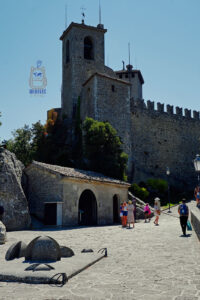 Średniowieczna twierdza Guaita, znana również jako La Rocca, w San Marino. Na skalistym wzgórzu wznosi się masywna kamienna wieża z dzwonnicą oraz wysokie mury obronne zwieńczone charakterystycznymi blankami. U podnóża twierdzy znajduje się niewielki kamienny budynek z łukowym wejściem. Na brukowanej ścieżce prowadzącej wzdłuż murów spacerują turyści. Nad zabytkową fortecą rozciąga się błękitne niebo.