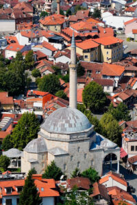 Widok z góry na zabytkowy meczet Emina Paszy w Prizren w Kosowie. Kamienna świątynia z dużą, szarą kopułą, mniejszymi kopułami i smukłym minaretem wyróżnia się pośród gęstej zabudowy historycznego centrum miasta. Otaczają ją tradycyjne domy z czerwonymi dachówkami, wąskie uliczki i zielone drzewa. Zdjęcie wykonane w słoneczny dzień ukazuje charakterystyczną architekturę osmańską oraz malowniczą panoramę Prizren.