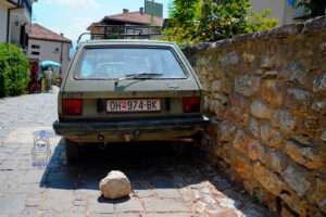 tara Zastava Yugo 45 w oliwkowozielonym kolorze, zaparkowana na wąskiej, brukowanej uliczce Starego Miasta w Ochrydzie w Macedonii Północnej. Samochód, będący jednym z symboli dawnej jugosłowiańskiej motoryzacji, ma charakterystyczną kanciastą sylwetkę, czarny zderzak, tylną wycieraczkę i metalowy bagażnik dachowy. Pojazd stoi tuż przy zabytkowym kamiennym murze, a za nim widoczne są tradycyjne domy z czerwonymi dachami. Na bruku za samochodem leży duży kamień. Zdjęcie oddaje nostalgiczny klimat bałkańskich uliczek, gdzie dawna motoryzacja wciąż jest częścią codziennego krajobrazu.