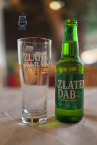 Zielona szklana butelka macedońskiego piwa Zlaten Dab stojąca obok pustej, przezroczystej szklanki z logo tej samej marki. Na etykiecie butelki widnieje duży biały napis „ZLATEN DAB” oraz oznaczenie „Macedonian Lager Beer”. Oba przedmioty ustawiono na jasnym obrusie, a rozmyte tło restauracji nadaje fotografii przytulny, wakacyjny charakter. Ciepłe światło podkreśla intensywną zieleń szkła i delikatne refleksy na szklance. Zdjęcie przedstawia jedną z popularnych marek piwa w Macedonii Północnej, będącą elementem lokalnych smaków poznawanych podczas podróży.