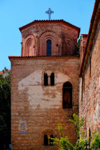 Zabytkowa cerkiew św. Zofii w Ochrydzie w Macedonii Północnej, przedstawiona z bliska w słoneczny dzień. Fotografia ukazuje fragment świątyni z charakterystyczną bizantyjską architekturą, wykonaną z czerwonej cegły i kamienia. Górną część budowli zdobią półkoliste ceglane łuki, wąskie okna oraz dach pokryty czerwoną dachówką, zwieńczony metalowym krzyżem. Na elewacji widoczne są dekoracyjne wzory ceglane, pozostałości jasnego tynku i ślady wielowiekowej historii budynku. Po prawej stronie znajduje się kamienno-ceglany mur sąsiedniej części świątyni, a u dołu kadru zielona roślinność. Intensywnie błękitne niebo podkreśla ciepłą kolorystykę zabytkowych murów. Zdjęcie eksponuje detale architektoniczne jednej z najważniejszych średniowiecznych świątyń Ochrydy.