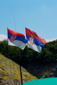 Dwie serbskie flagi w czerwono-niebiesko-białych barwach, z godłem przedstawiającym dwugłowego orła, powiewają na wietrze. W tle zielone wzgórza, skaliste zbocza i błękitne niebo