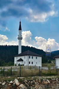 Niewielki, biały meczet z ciemnym, czterospadowym dachem i wysokim, smukłym minaretem zakończonym czarną iglicą, położony w górskiej miejscowości w Czarnogórze. Budynek otaczają kamienne murki i metalowe ogrodzenie, a w tle wznoszą się wzgórza porośnięte gęstym lasem iglastym. Nad meczetem rozciąga się błękitne niebo z imponującymi białymi i szarymi chmurami. Zdjęcie ukazuje charakterystyczne połączenie lokalnej architektury sakralnej z malowniczym krajobrazem czarnogórskiej prowincji.