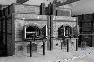 Czarno-biała fotografia przedstawiająca historyczne piece krematoryjne w budynku krematorium byłego nazistowskiego obozu koncentracyjnego Dachau w Niemczech. Na zdjęciu widoczne są dwa murowane z cegły piece z otwartymi metalowymi drzwiczkami oraz wysuniętymi szynami służącymi do wprowadzania ciał do komór spalania. Surowe wnętrze, masywne konstrukcje pieców i monochromatyczna kolorystyka podkreślają tragiczny charakter tego miejsca, będącego świadectwem zbrodni nazistowskich i cierpienia więźniów obozu.
