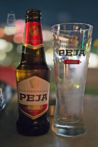 Butelka kosowskiego piwa Birra Peja Pilsner stojąca obok wysokiej, pustej szklanki z logo tego samego browaru. Ciemna szklana butelka z charakterystyczną czerwono-kremową etykietą pokryta jest kroplami wilgoci, a na schłodzonej szklance widoczna jest para wodna. W tle rozmyte, kolorowe światła wieczornego miasta tworzą przyjemną atmosferę. Zdjęcie przedstawia popularne lokalne piwo z browaru Peja, będące jednym z rozpoznawalnych produktów Kosowa.
