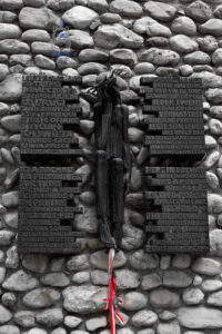 Fotografia przedstawiająca pomnik Chrystusa Frasobliwego na terenie miejsca pamięci byłego nazistowskiego obozu koncentracyjnego Dachau w Niemczech. Na tle kamiennego muru znajduje się ciemna rzeźba cierpiącego Chrystusa w koronie cierniowej, otoczona czterema tablicami pamiątkowymi z napisami w językach polskim, niemieckim, francuskim i angielskim. Tablice przypominają o polskich duchownych więzionych i zamordowanych w Dachau. U stóp rzeźby umieszczono biało-czerwone wstążki, symbolizujące pamięć o ofiarach. Czarno-biała kolorystyka fotografii, z wyeksponowanymi barwami polskiej flagi, podkreśla powagę i symboliczne znaczenie tego miejsca.