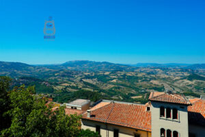 Panoramiczny widok z góry Monte Titano w San Marino na rozległe wzgórza i doliny Apeninów. Na pierwszym planie widoczne są budynki z charakterystycznymi czerwonymi dachówkami, otoczone bujną zielenią. W oddali rozciąga się malowniczy krajobraz zielonych lasów, złocistych pól uprawnych i łagodnych górskich grzbietów. Całość dopełnia intensywnie błękitne, bezchmurne niebo, podkreślające słoneczną atmosferę letniego dnia.