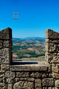 Panoramiczny widok z murów obronnych średniowiecznej wieży Guaita w San Marino. Przez otwór pomiędzy masywnymi kamiennymi blankami rozciąga się krajobraz łagodnych wzgórz, zielonych dolin, pól uprawnych i niewielkich skupisk zabudowań. Na horyzoncie widoczne są kolejne pasma wzgórz, a nad nimi rozpościera się intensywnie błękitne, bezchmurne niebo. Surowe kamienne mury twierdzy tworzą naturalną ramę dla rozległej panoramy.